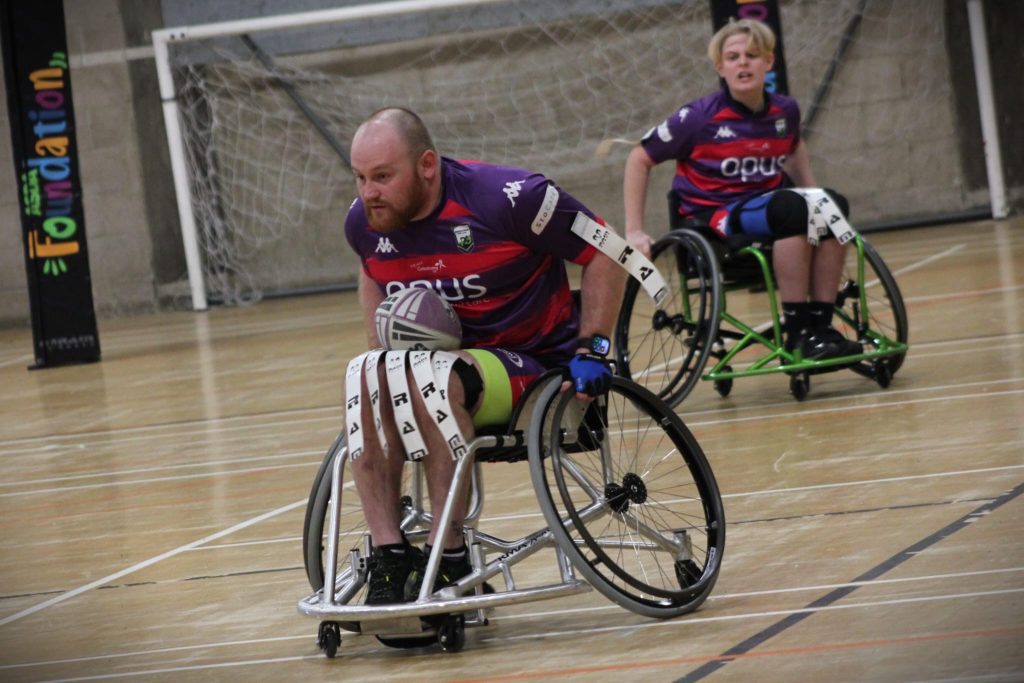A Team colostomy UK wheelchair rugby league player is wheeling across the court with a rugby ball in his lap.