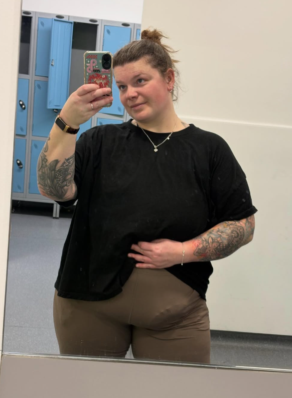 Emily is taking a mirror selfie with gym lockers in the background and her stoma bag slightly visible through her leggings.