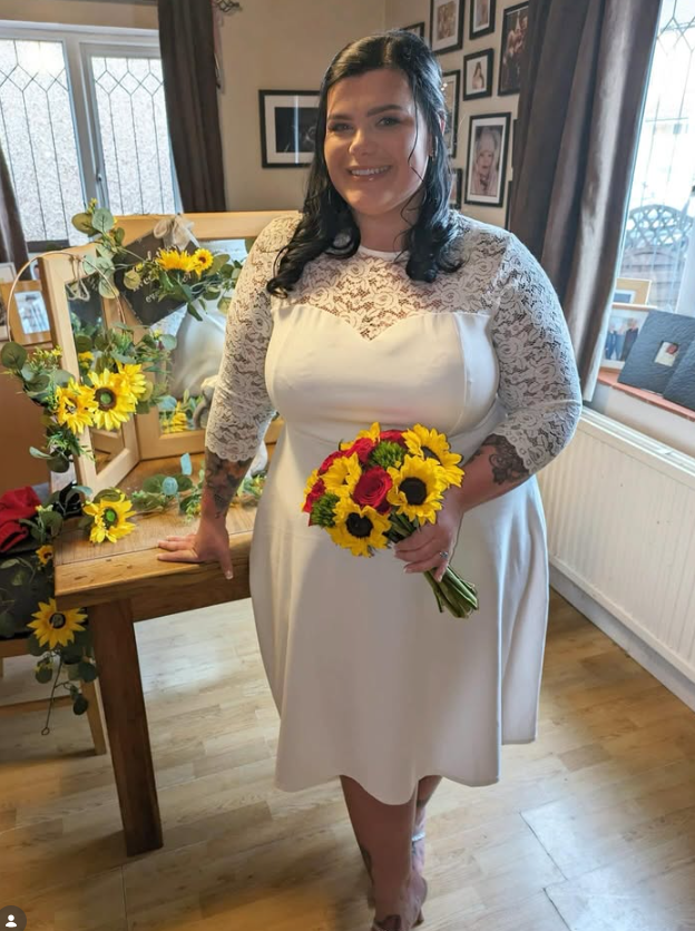 Emily smiles at the camera wearing a white wedding dress, holding a bouquet of sunflowers and roses.