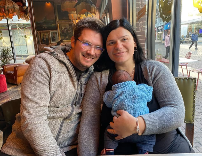 Emily is holding her new born son and smiling at the camera, Emily's husband is sat close to her, also smiling.