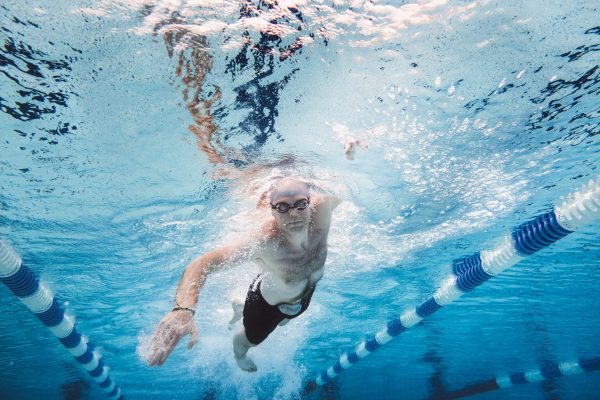 Full length of shirtless sportsman practicing freestyle swimming in pool. Mid adult male athlete is training underwater amidst lane marker. Image represents healthy lifestyle.